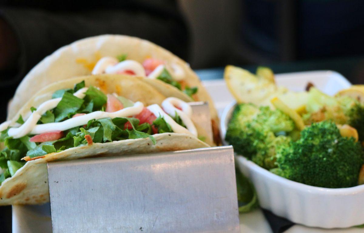 Two tacos sit on a taco holder with lettuce, tomato and sour cream on top. Next to the tacos is a small bowl of cooked vegetables.