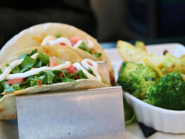 Two tacos sit on a taco holder with lettuce, tomato and sour cream on top. Next to the tacos is a small bowl of cooked vegetables.