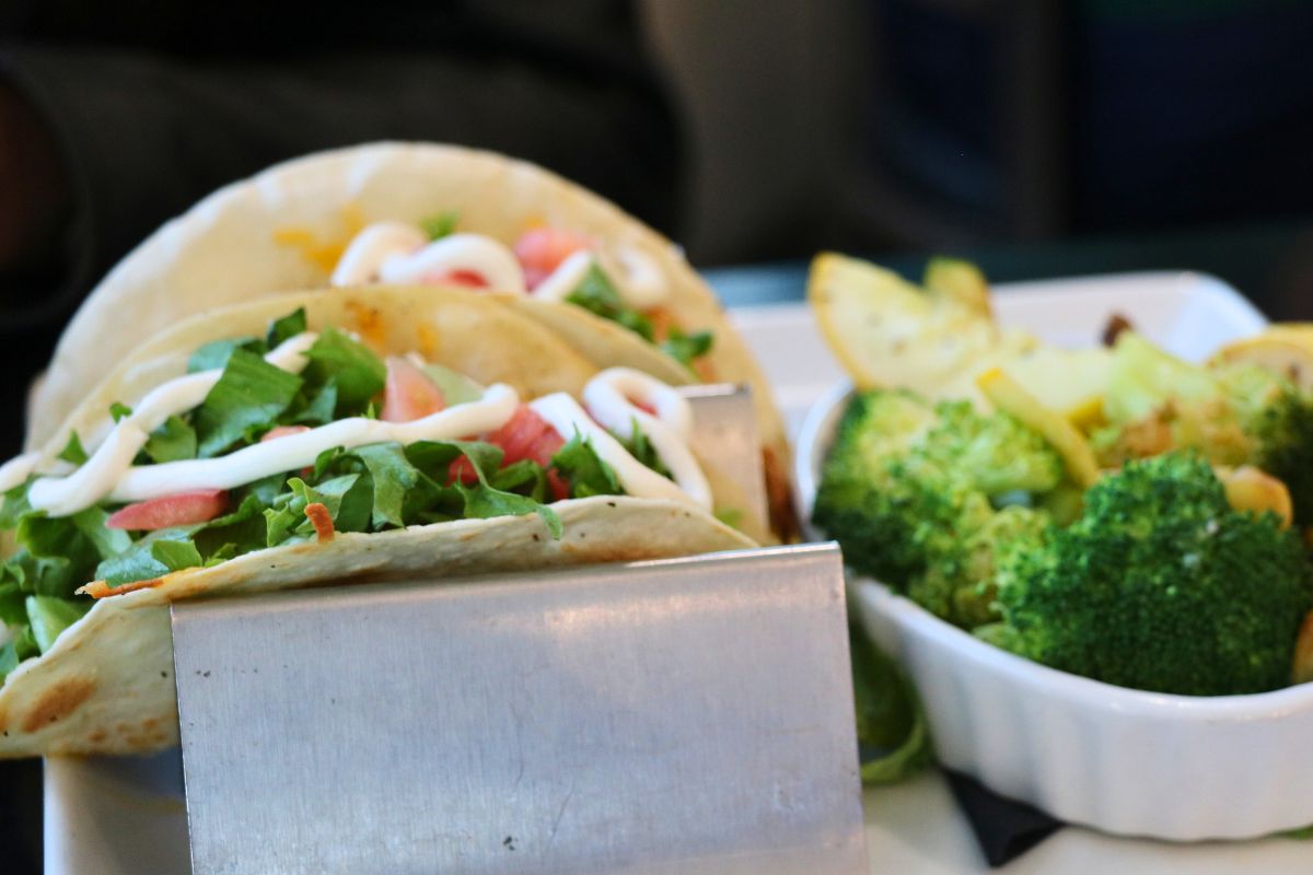Two tacos sit on a taco holder with lettuce, tomato and sour cream on top. Next to the tacos is a small bowl of cooked vegetables.