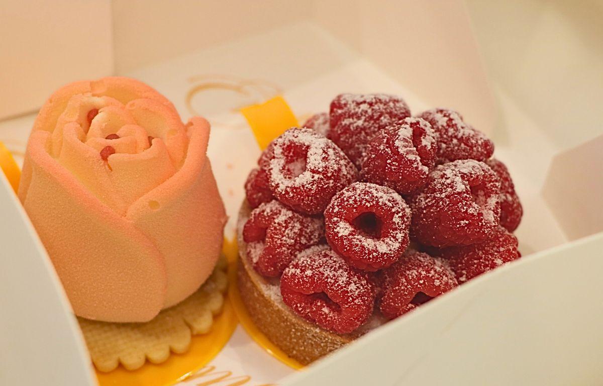 Two tarts sit next to each other in a white box. One tart features raspberries displayed in circles with a white powder on top. Underneath is a bread base and a yellow dish to hold the desert. The other pastry is pink and rose shaped with a rose cream poking from the top. Underneath the rose is a cracker on top of a yellow dish to hold the pastry.