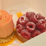 Two tarts sit next to each other in a white box. One tart features raspberries displayed in circles with a white powder on top. Underneath is a bread base and a yellow dish to hold the desert. The other pastry is pink and rose shaped with a rose cream poking from the top. Underneath the rose is a cracker on top of a yellow dish to hold the pastry.