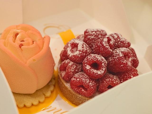 Two tarts sit next to each other in a white box. One tart features raspberries displayed in circles with a white powder on top. Underneath is a bread base and a yellow dish to hold the desert. The other pastry is pink and rose shaped with a rose cream poking from the top. Underneath the rose is a cracker on top of a yellow dish to hold the pastry.