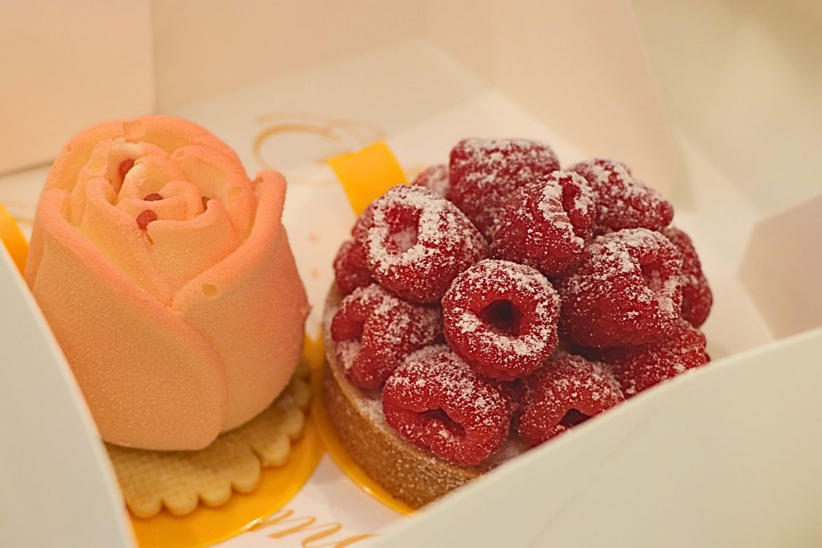 Two tarts sit next to each other in a white box. One tart features raspberries displayed in circles with a white powder on top. Underneath is a bread base and a yellow dish to hold the desert. The other pastry is pink and rose shaped with a rose cream poking from the top. Underneath the rose is a cracker on top of a yellow dish to hold the pastry.