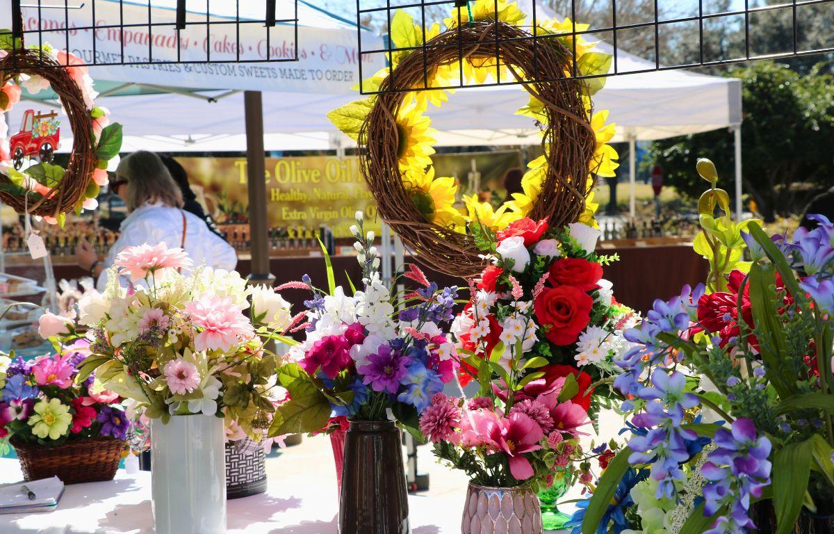 Flowers are displayed on a table under a canopy with a market in the background.