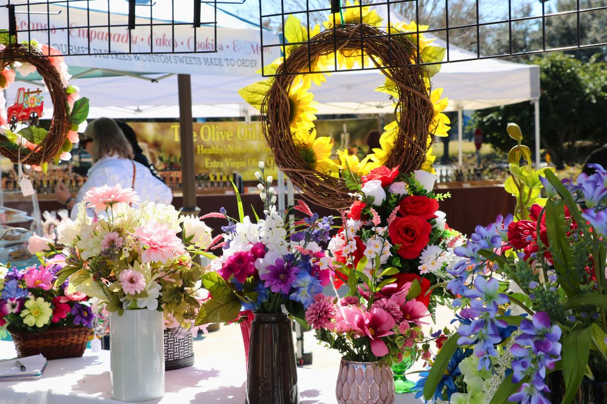 Flowers are displayed on a table under a canopy with a market in the background.