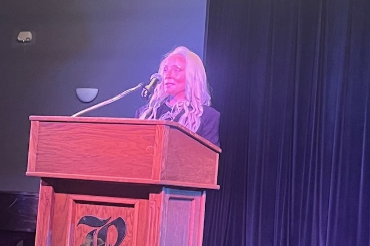 A woman standing behind a podium with an insignia, with a microphone in front of her face, with a dark wall and curtain in the background.