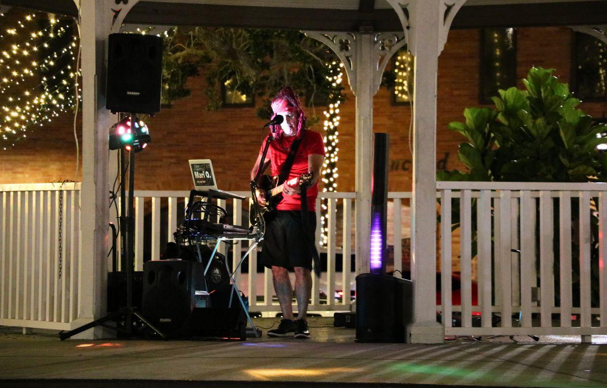A performer dressed in a red shirt, bandana and black shorts stands behind a microphone with a guitar with various instruments and speakers around him.