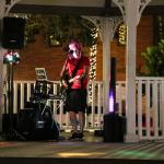 A performer dressed in a red shirt, bandana and black shorts stands behind a microphone with a guitar with various instruments and speakers around him.