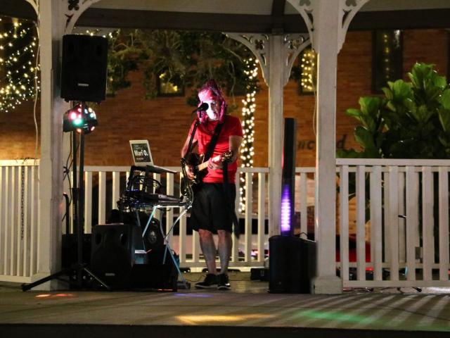 A performer dressed in a red shirt, bandana and black shorts stands behind a microphone with a guitar with various instruments and speakers around him.