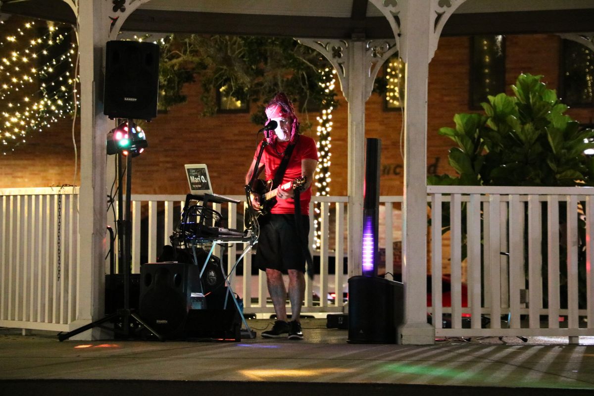 A performer dressed in a red shirt, bandana and black shorts stands behind a microphone with a guitar with various instruments and speakers around him.