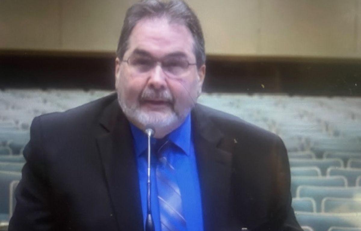 A man dressed in a suit standing before a microphone with gray seats, a brown baseboard and beige wall int he background.