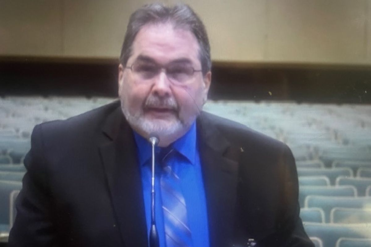 A man dressed in a suit standing before a microphone with gray seats, a brown baseboard and beige wall int he background.