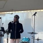 A drum kit in the background against a tarp wall on a stage, with a woman in the foreground singing behind a microphone, with microphones to her right and left.
