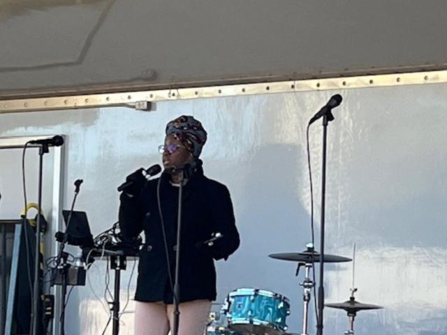 A drum kit in the background against a tarp wall on a stage, with a woman in the foreground singing behind a microphone, with microphones to her right and left.