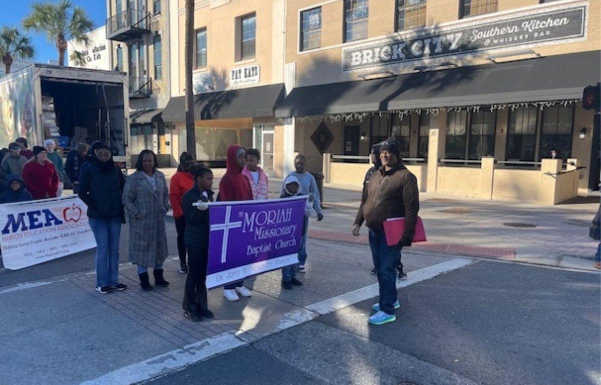 A group of people standing behind a banner on a street in front of store fronts, with a man on the extreme right standing in front of the banner.