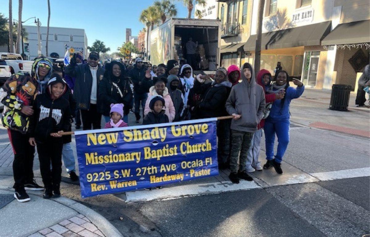 A truck, store front and palm trees in the background. A group of people standing behind a blue banner with writing on it.