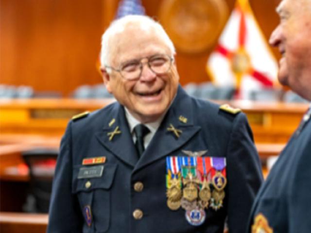 A man dressed in a miliary uniform, with a slew of decorations on the right side of his chest, campaign ribbons on the left, with man to his right. In the background, seats, in the far background, a wood grain wall, and a flag of the State of Florida to the right.