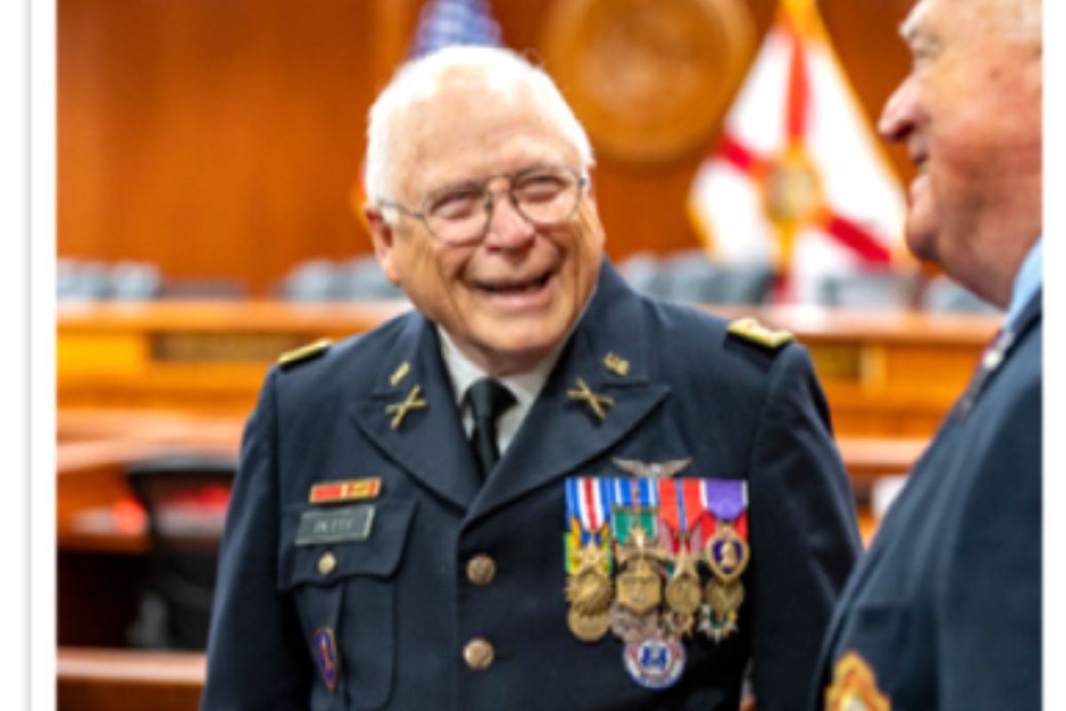 A man dressed in a miliary uniform, with a slew of decorations on the right side of his chest, campaign ribbons on the left, with man to his right. In the background, seats, in the far background, a wood grain wall, and a flag of the State of Florida to the right.