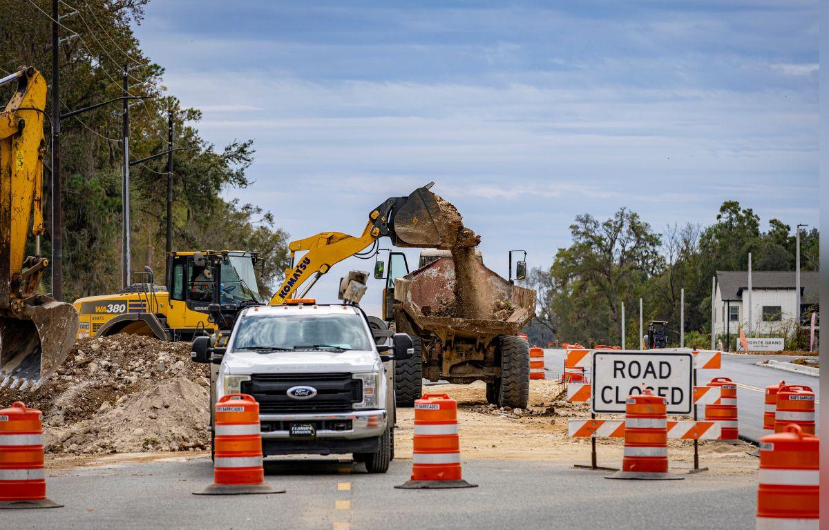 A yellow backhoe, a white pickup truck with orange and white traffic control barricades in front of it to the right. Behind the vehicle to the left, a pile of dirt, behind the vehicle in the background, a trackhoe, trees in the background to the extreme left and right, a traffic control sign with writing to the right. in the foreground paved road.