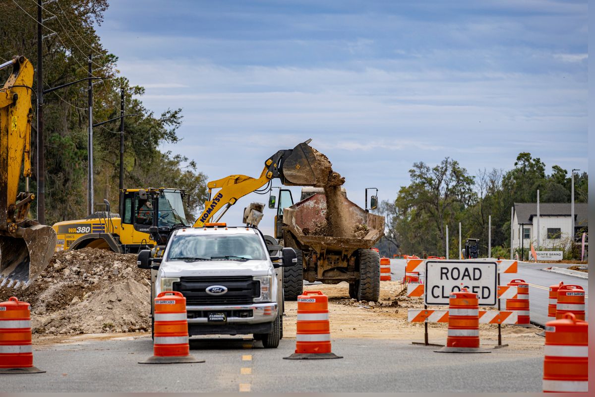 A yellow backhoe, a white pickup truck with orange and white traffic control barricades in front of it to the right. Behind the vehicle to the left, a pile of dirt, behind the vehicle in the background, a trackhoe, trees in the background to the extreme left and right, a traffic control sign with writing to the right. in the foreground paved road.