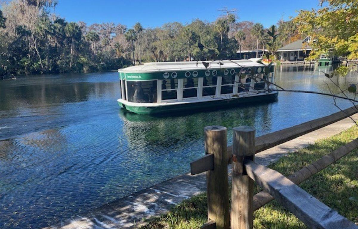 In the background a tree line and buildings. A vessel on a waterway, with sidewalk, grass and paddock fencing in the foreground.