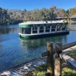In the background a tree line and buildings. A vessel on a waterway, with sidewalk, grass and paddock fencing in the foreground.