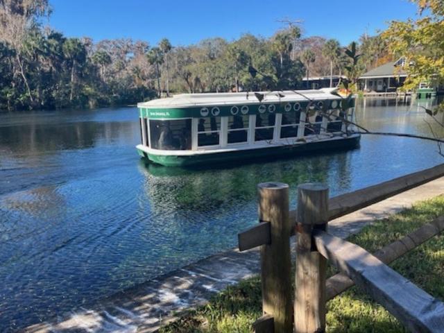 In the background a tree line and buildings. A vessel on a waterway, with sidewalk, grass and paddock fencing in the foreground.
