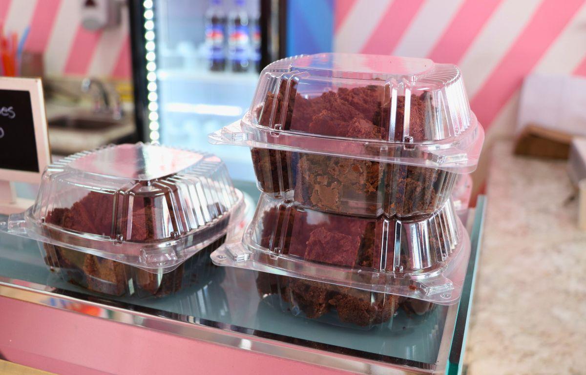 Brownies in plastic containers sit on a counter with a drink cooler in the background against pink and white diagonally striped walls.