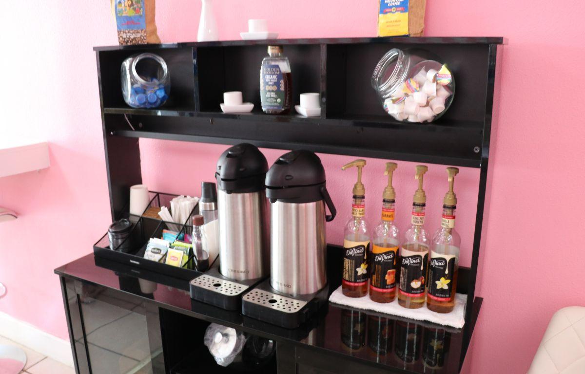 A black shelf in front of a pink wall features items to make coffee including coffee machines, creamers, cups, sugar and more.