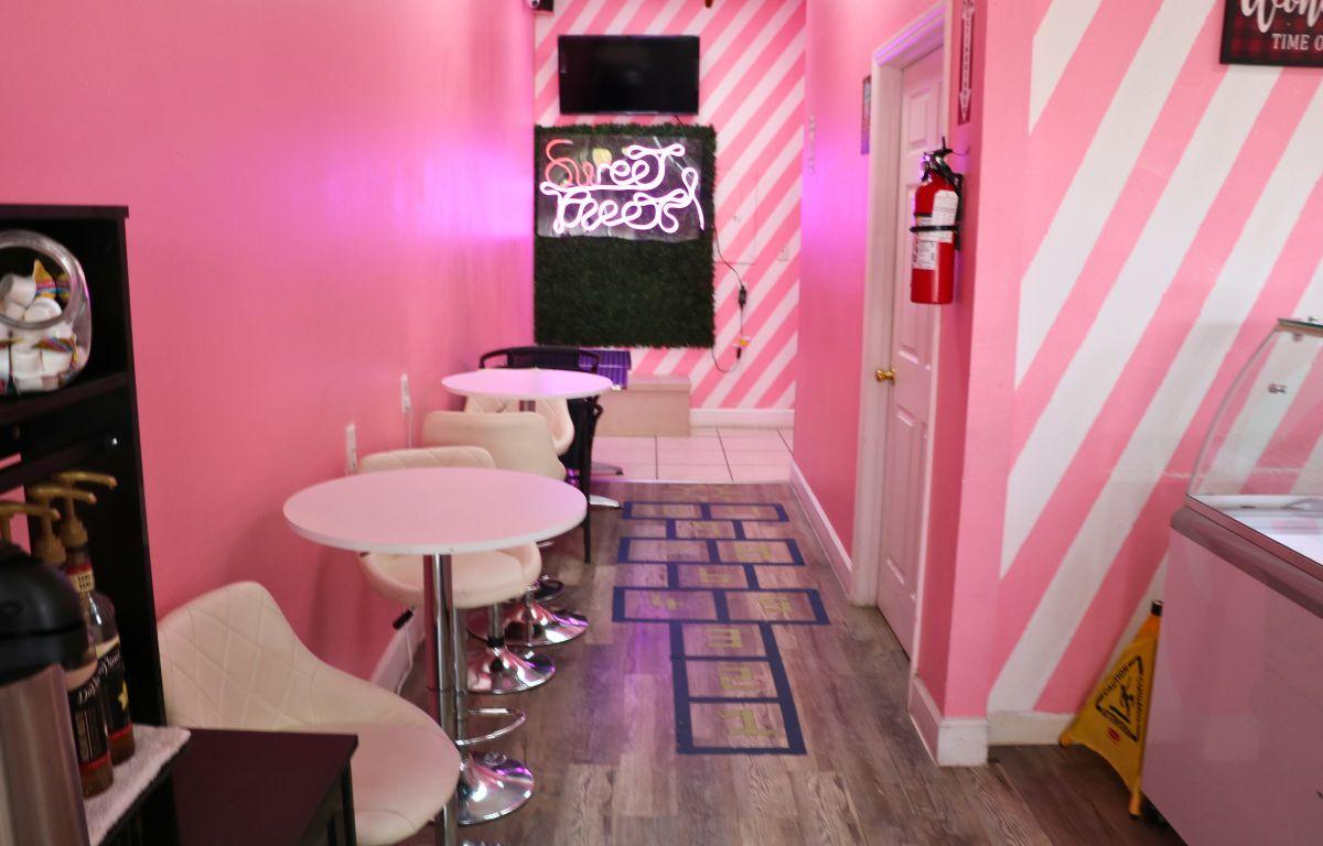 A store with pink and white walls features wood floors with a black hopscotch outline, white tables and bar stools and a neon light sign that reads, "Sweet Treets."