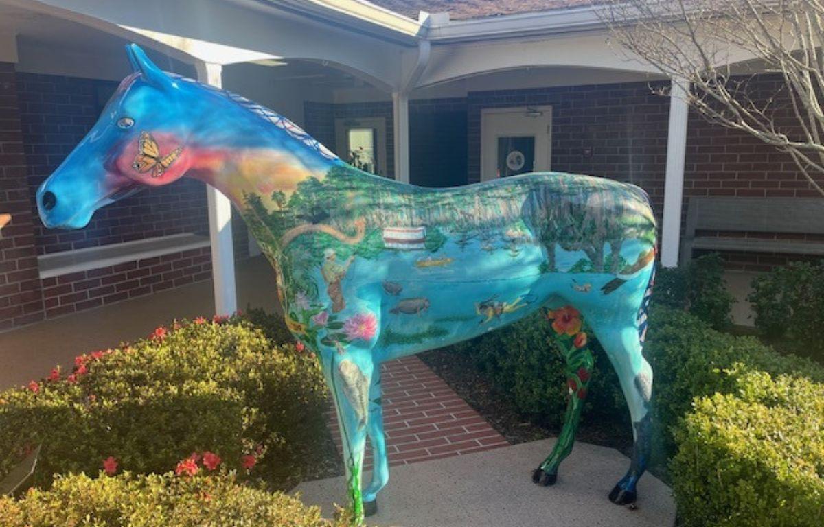 Shrubbery on the left, a fiberglass horse in the foreground on a concrete platform, trim and columns in the background, and in the far background, brick, windows and doors.