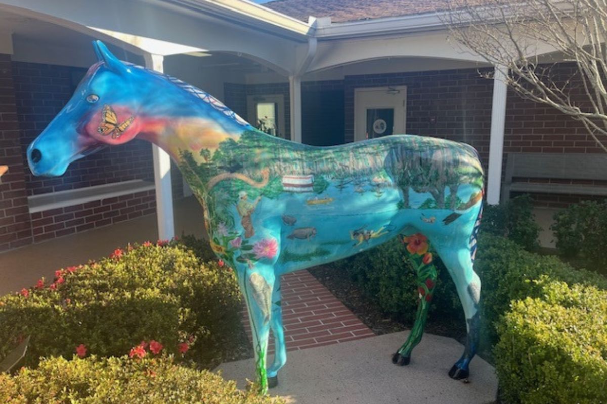 Shrubbery on the left, a fiberglass horse in the foreground on a concrete platform, trim and columns in the background, and in the far background, brick, windows and doors.