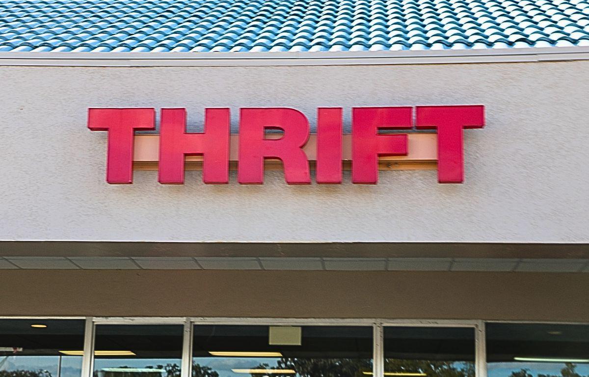 A storefront features a blue roof with beige walls. A large, red sign reads, "Thrift." A wall of glass windows and a door lie just below the sign.