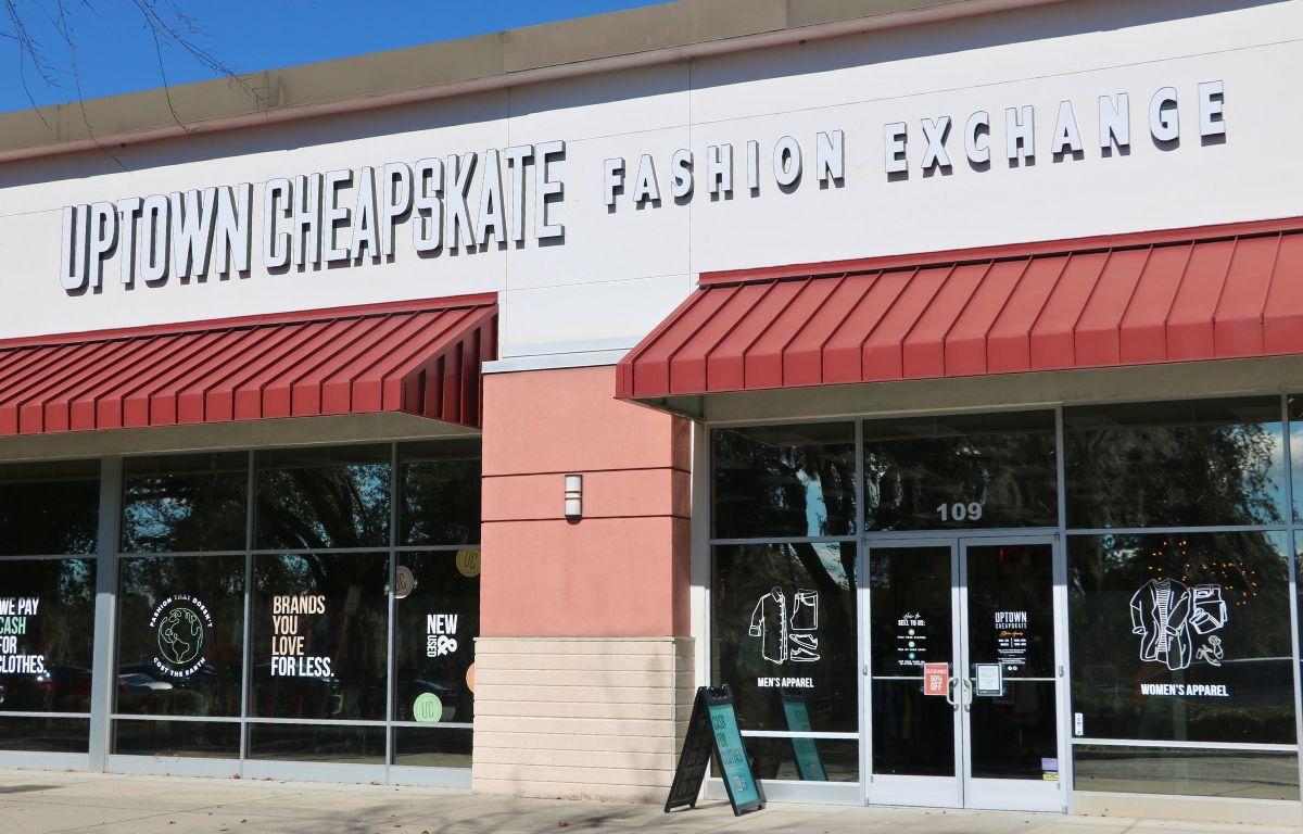 A storefront features beige, peach and white walls with red overhangs. under the overhangs are glass walls with glass doors. A sidewalk is in front of the stores. On the white section above the overhangs is a large, white, bold sign reading, "Uptown Cheapstake Fashion Exchange."