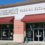 A storefront features beige, peach and white walls with red overhangs. under the overhangs are glass walls with glass doors. A sidewalk is in front of the stores. On the white section above the overhangs is a large, white, bold sign reading, "Uptown Cheapstake Fashion Exchange."