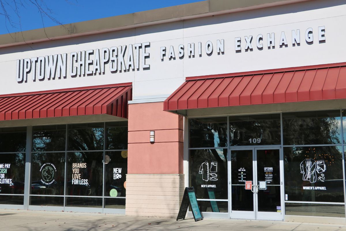 A storefront features beige, peach and white walls with red overhangs. under the overhangs are glass walls with glass doors. A sidewalk is in front of the stores. On the white section above the overhangs is a large, white, bold sign reading, "Uptown Cheapstake Fashion Exchange."