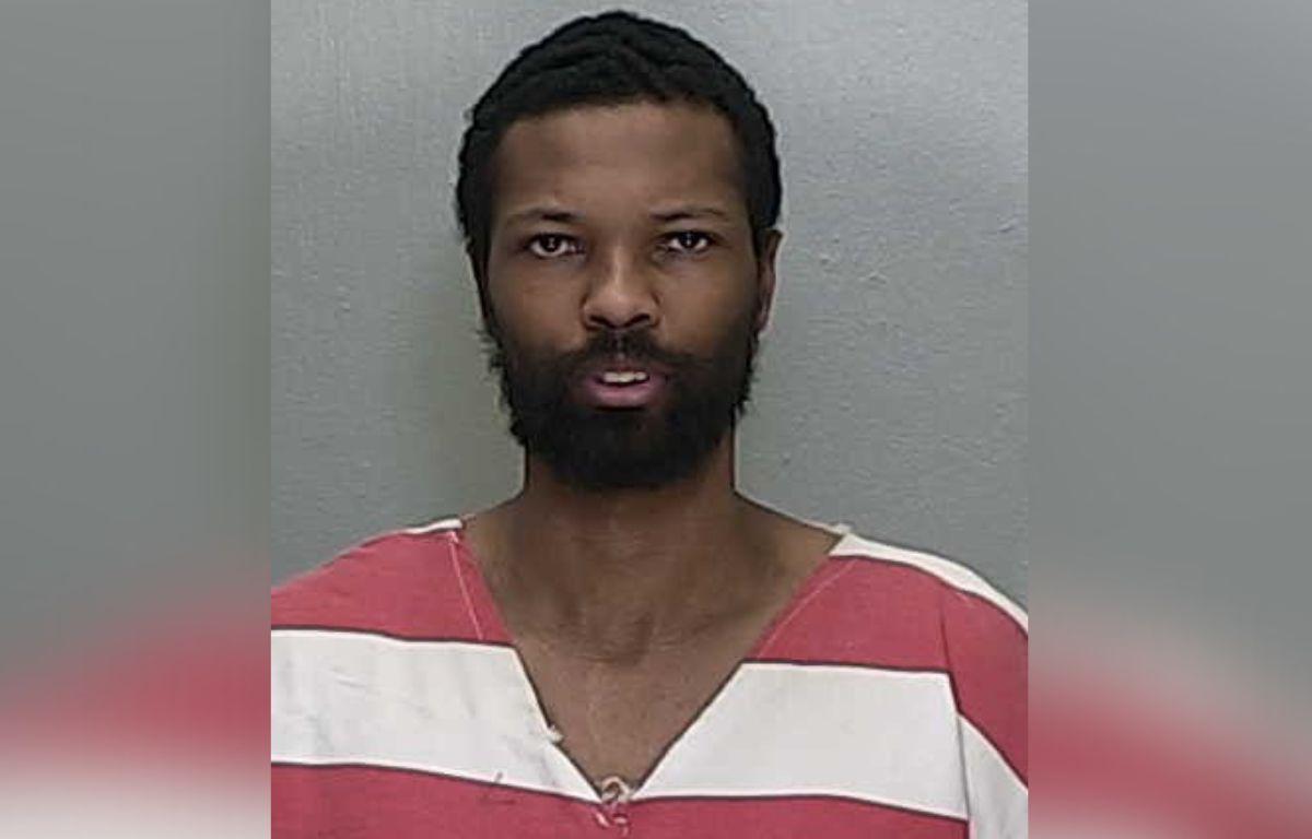 A man in an orange and white jump suit standing against a gray wall.