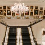 A grand staircase leads up to a second floor of a large, white manor. Paintings of dogs line the wall behind the stairs on the second floor. A large chandelier hangs above the staircase.