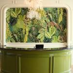 An entrance to a restaurant features wallpaper with vibrant, green plants on them. A green, semi-circle hostess stand with a small device behind it stands in the center of the wall. A chandelier with white, faux flowers on it hangs above the hostess stand.