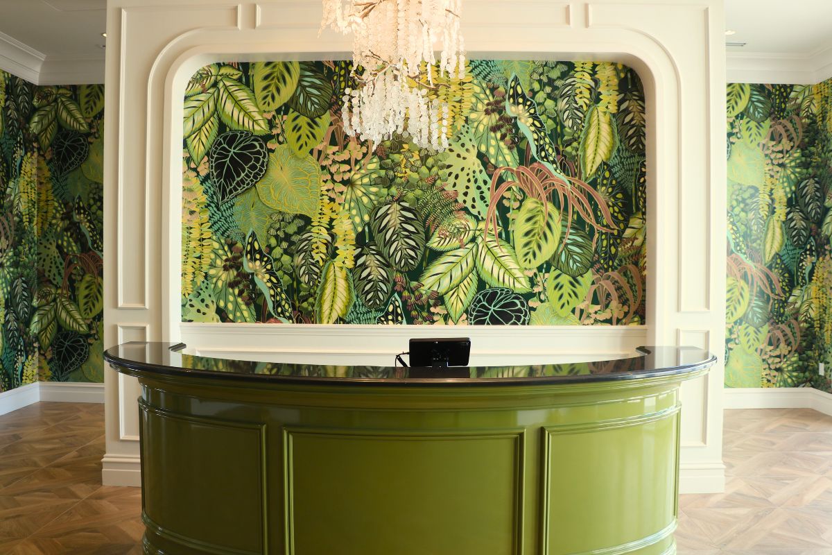 An entrance to a restaurant features wallpaper with vibrant, green plants on them. A green, semi-circle hostess stand with a small device behind it stands in the center of the wall. A chandelier with white, faux flowers on it hangs above the hostess stand.