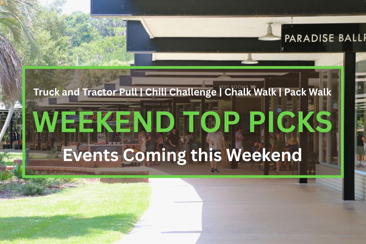 People walk and gather in a shaded outdoor shopping and dining area. Signs above indicate locations such as "Paradise Ballroom," "Rest Rooms," "Snack Shop," "Springside Café," and "Discovery Center." A food cart selling pretzels is positioned along the walkway, and some visitors are seated at tables near storefronts. The area is surrounded by green grass and trees. Green and white text inside the box reads, "Truck and Tractor Pull | Chili Challenge | Chalk Walk | Pack Walk. WEEKEND TOP PICKS. Events Coming this Weekend."