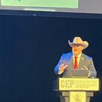 A black background, with a screen overhead, a man in a suit and cowboy hat gesticulating with his hands, in front of a podium with placards, that have writing and insignias, with the podium having a microphone.