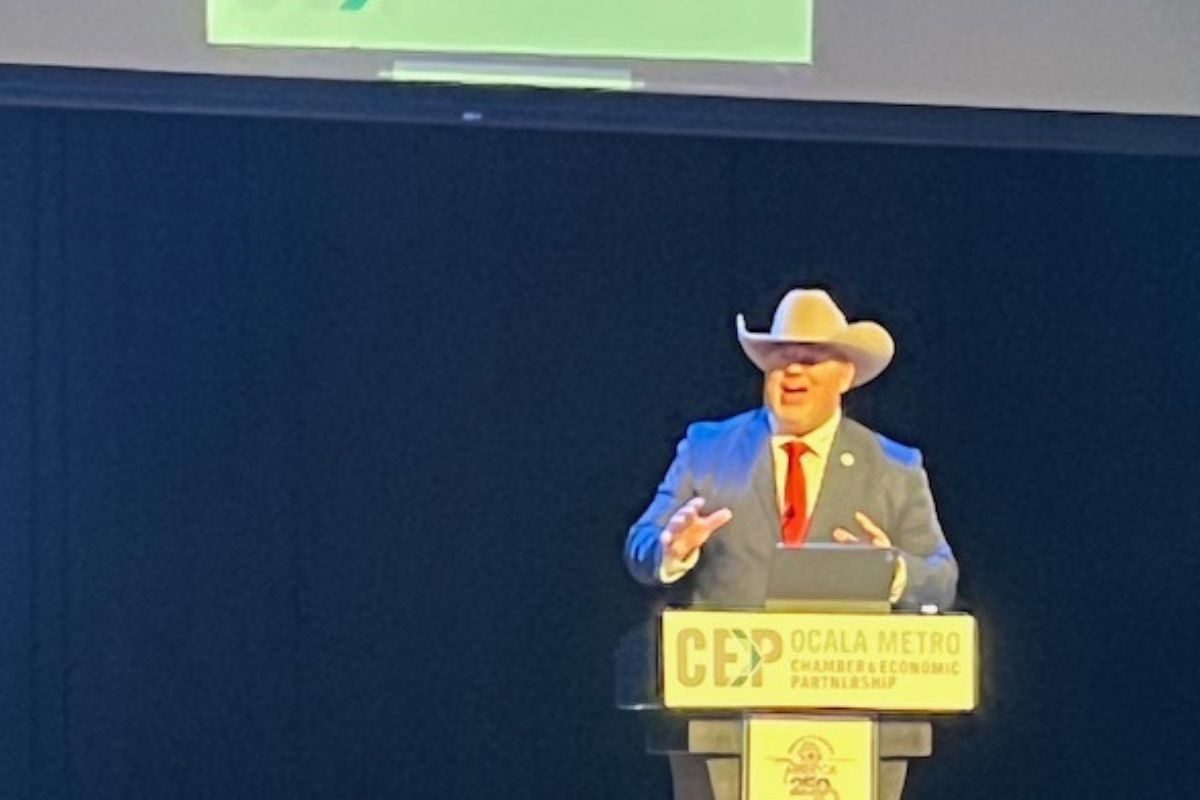 A black background, with a screen overhead, a man in a suit and cowboy hat gesticulating with his hands, in front of a podium with placards, that have writing and insignias, with the podium having a microphone.