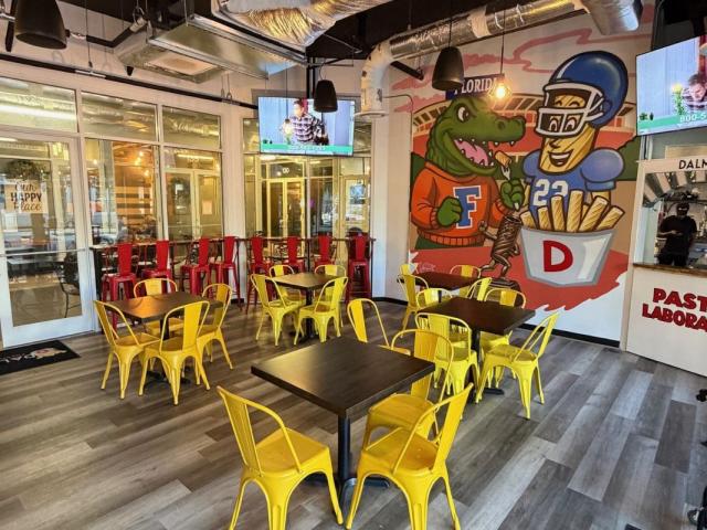 A colorful dining space with yellow metal chairs and wooden tables. A mural of an alligator in a Florida Gators jersey posing with a football player sits in the background.