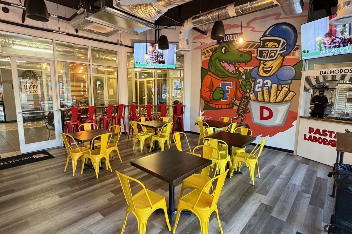 A colorful dining space with yellow metal chairs and wooden tables. A mural of an alligator in a Florida Gators jersey posing with a football player sits in the background.