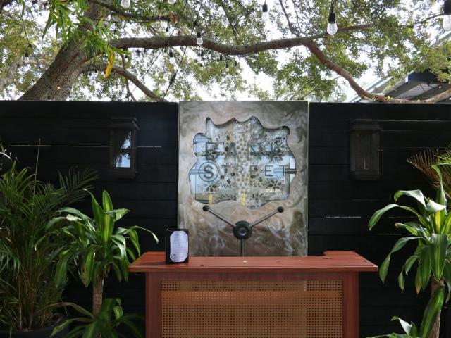 The entrance of a restaurant features a wooden hostess stand in front of a metal panel with text that reads, "Bank Street." The panel is against a black, wooden wall with plants on either side of the hostess stand.