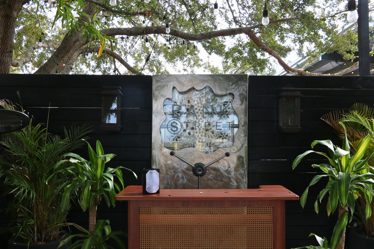 The entrance of a restaurant features a wooden hostess stand in front of a metal panel with text that reads, "Bank Street." The panel is against a black, wooden wall with plants on either side of the hostess stand.