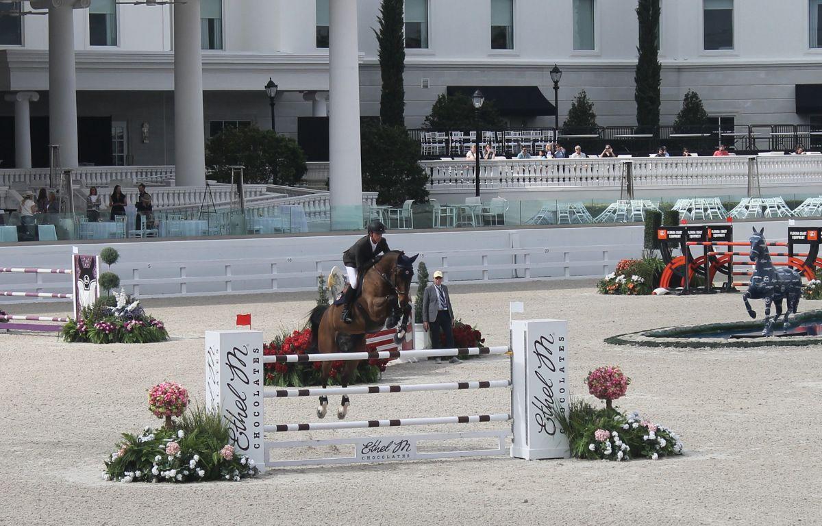 A horse and rider jumping over a vertical jumping effort, in an arena, with columns, seating, lights, ornamental vegetation, windows in front of a building. There are jumping efforts to the left and right, and a fiberglass horse to the right.