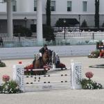 A horse and rider jumping over a vertical jumping effort, in an arena, with columns, seating, lights, ornamental vegetation, windows in front of a building. There are jumping efforts to the left and right, and a fiberglass horse to the right.