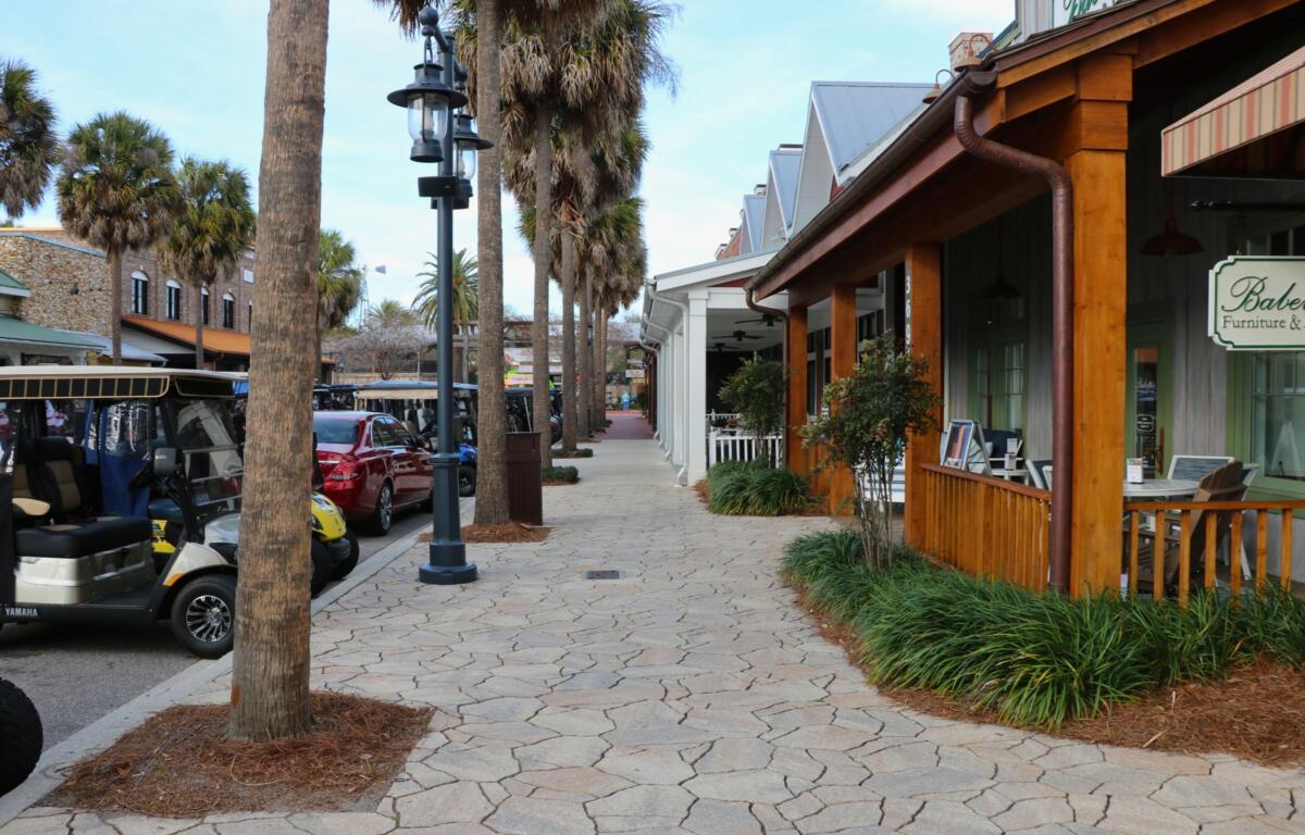 A sidewalk features shops and palm trees lined along it. Golf carts and cars are parked on the road next to the sidewalk.
